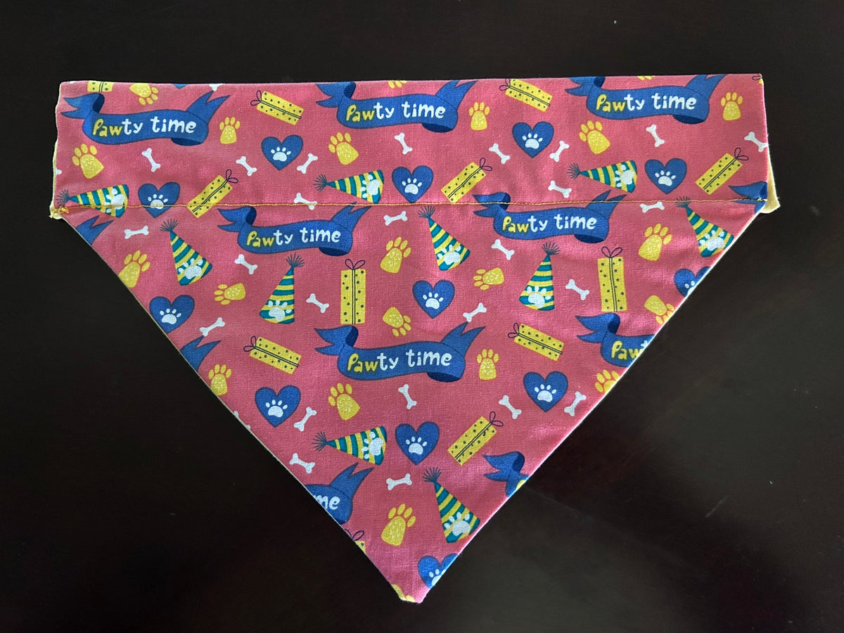 A triangular pet bandana with a pink/orange tinted background and a colorful 'Pawty time' pattern featuring blue hearts, yellow paw prints, and green-and-yellow gift packages, laid flat on a dark surface.