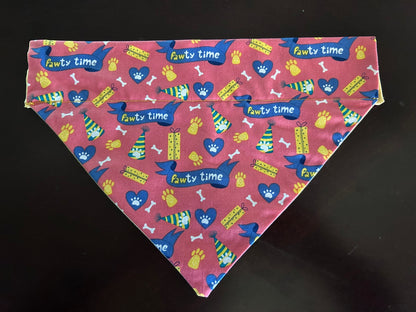 A triangular pet bandana with a pink/orange tinted background and a colorful 'Pawty time' pattern featuring blue hearts, yellow paw prints, and green-and-yellow gift packages, laid flat on a dark surface.