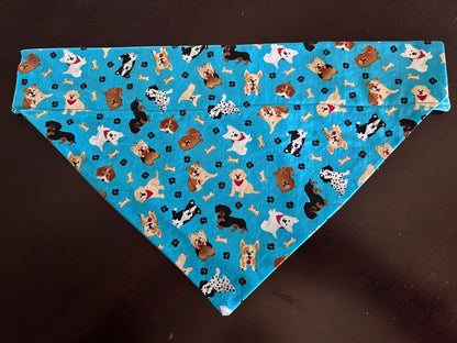 A triangular pet bandana with a bright blue background, printed with a pattern of various dog breeds and small black paw prints, laid flat on a dark brown surface.