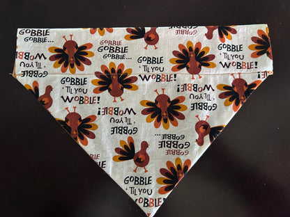 Triangular bandana lying on a wood surface.  Fabric is a cream background with turkeys and Gobble 'Til You Wobble text.