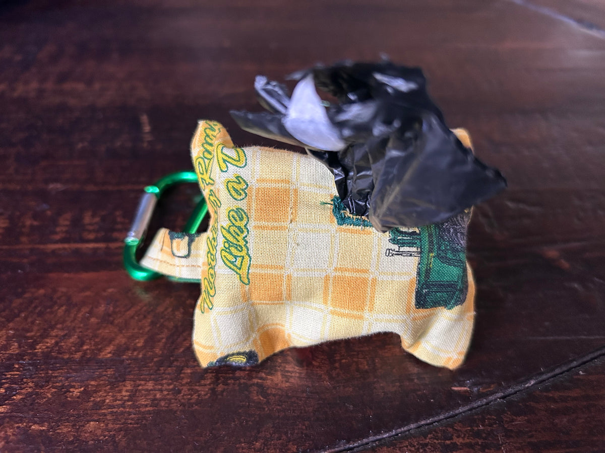 Waste Bag holder made of John Deer Fabric wth yellow checked background and green tractors holding a roll of black plastic bags lying on a wood surface.