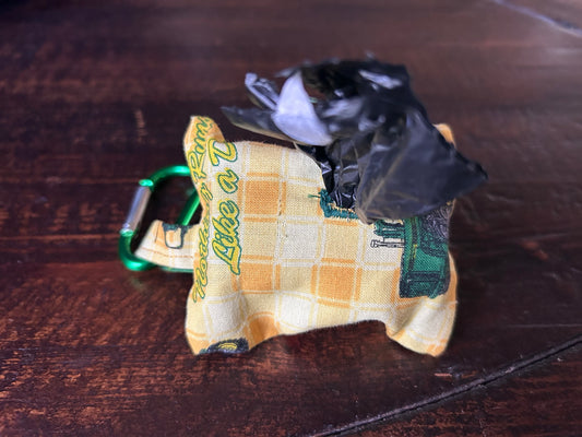 Waste Bag holder made of John Deer Fabric wth yellow checked background and green tractors holding a roll of black plastic bags lying on a wood surface.