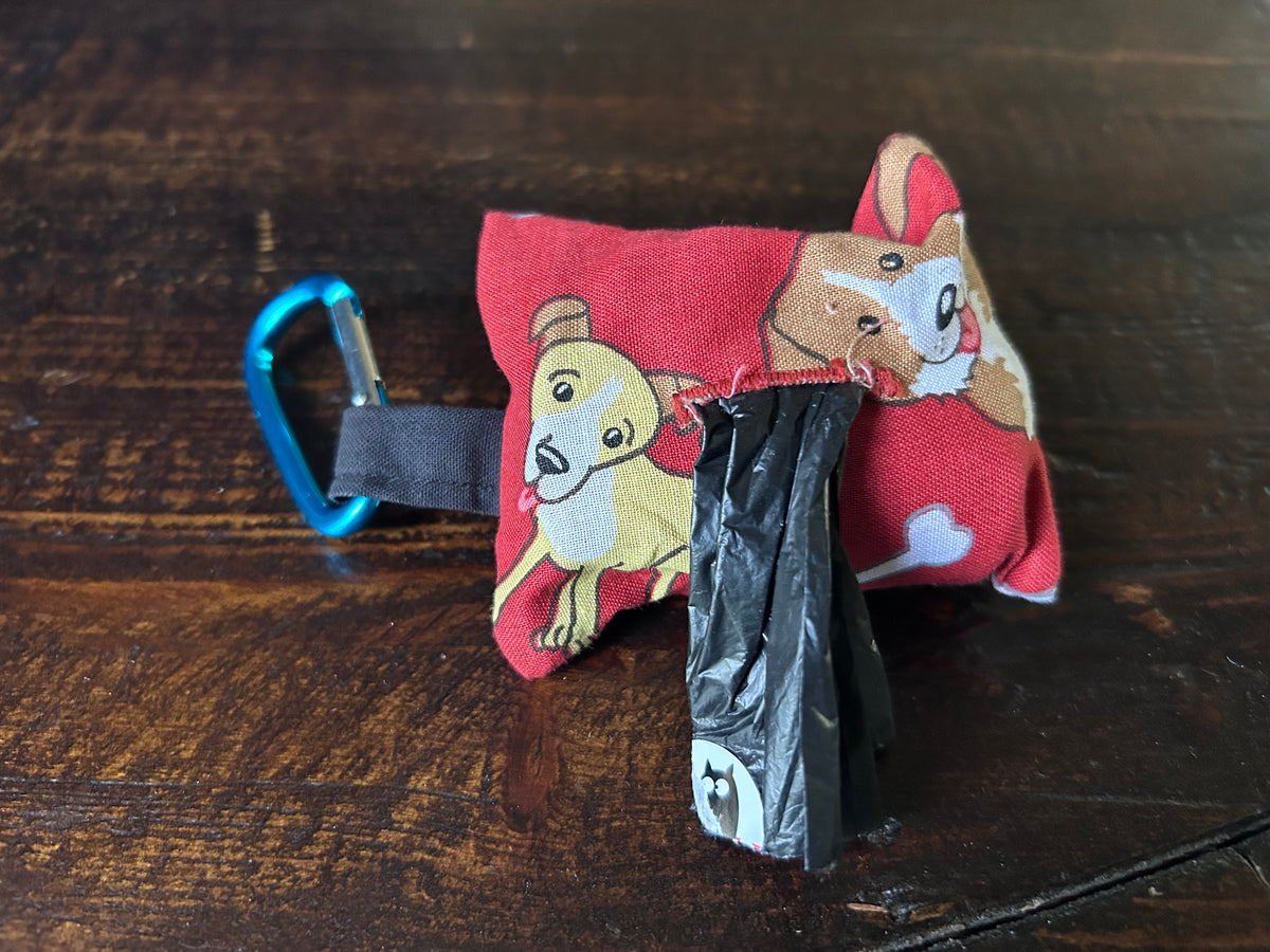 A small red fabric dog waste bag with cartoon dog prints, attached to a blue carabiner and holding a roll of black plastic bags, placed on a dark wooden surface.