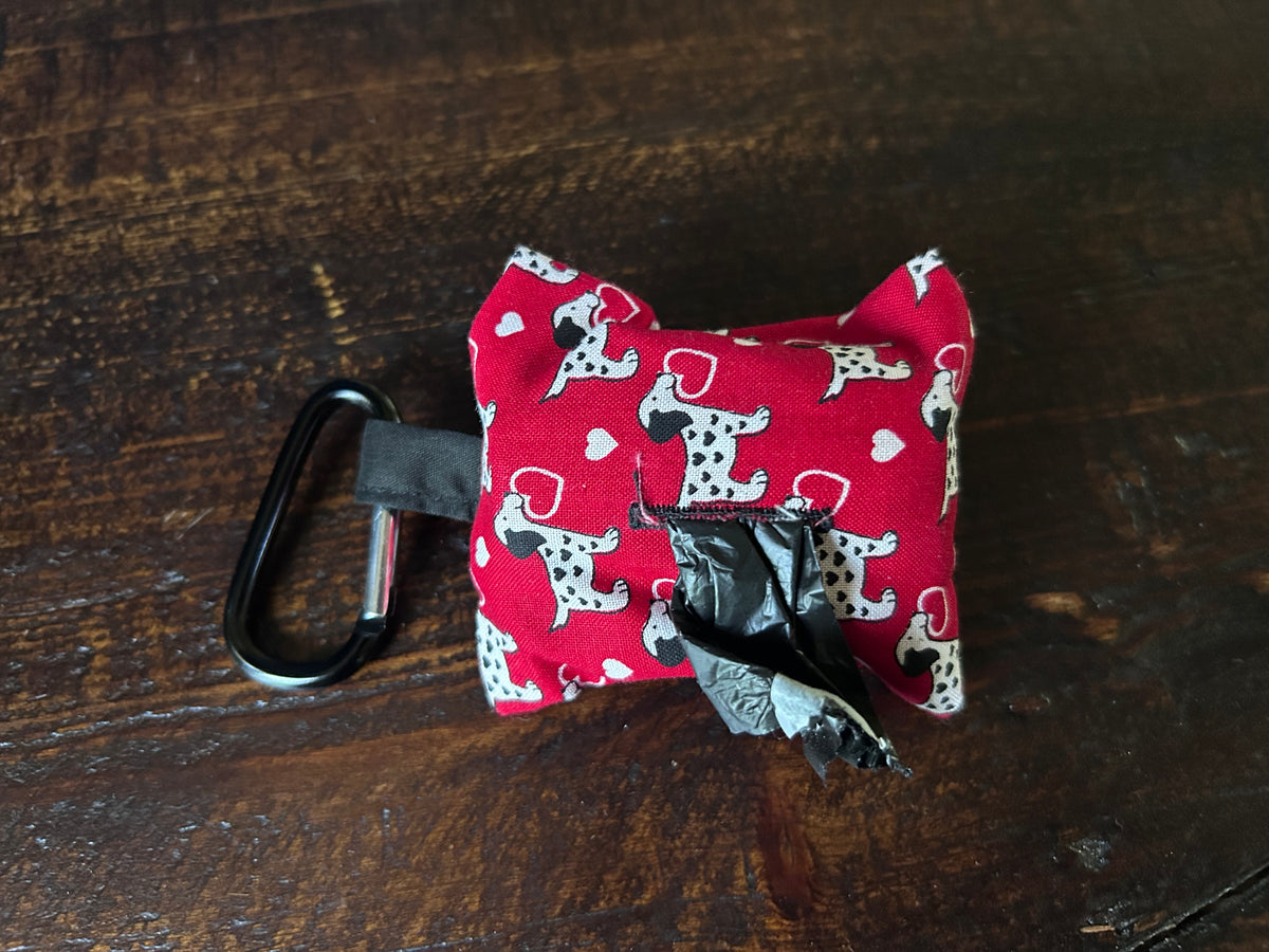 A small red fabric dog waste bag with white dalmation printed fabric, attached to a black carabiner and holding a roll of  black plastic bags, placed on a dark wooden surface.
