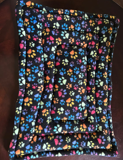 Rectangular pet bed made of black fleece fabric with a colorful rainbow paw print pattern, shown on a dark wooden surface.