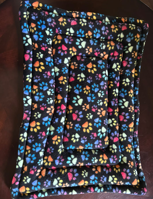Rectangular pet bed made of black fleece fabric with a colorful rainbow paw print pattern, shown on a dark wooden surface.