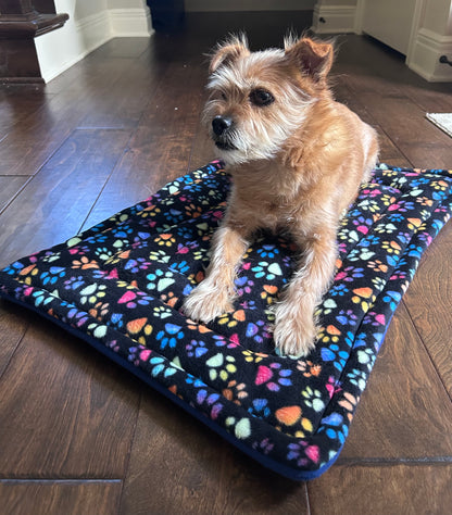A small tan dog sits on a rectangular pet mat with a black background and colorful paw-print pattern, placed on a dark wood floor in an indoor room.